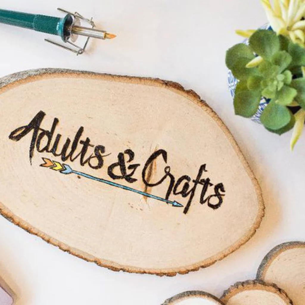 Adults and Crafts branding on a wooden craft panel beside a wood-burning tool.